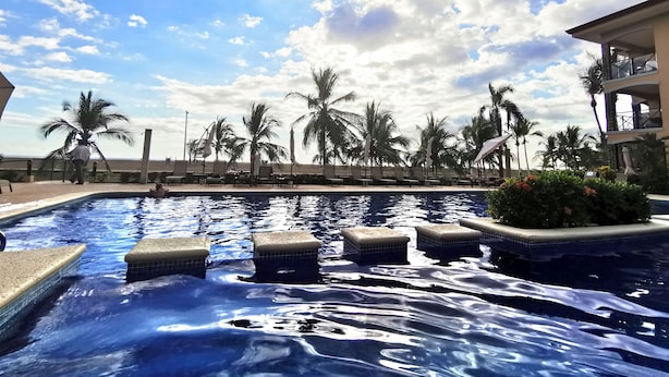 A swimming pool with clear water, surrounded by palm trees and lounge chairs, under a partly cloudy sky. - Beachfront Jaco Condos - Fully Equipped - 6 Pax (Jacó)