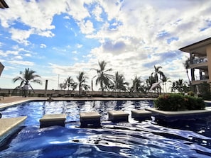 A swimming pool with clear water, surrounded by palm trees and lounge chairs, under a partly cloudy sky. - Beachfront Jaco Condos - Fully Equipped - 6 Pax (Jacó)