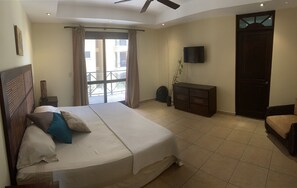 A bedroom with a large bed, a ceiling fan, a TV, a wooden door, and a view through a window. - Beachfront Jaco Condos - Fully Equipped - 6 Pax (Jacó)