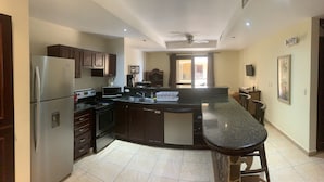 A modern kitchen with dark wooden cabinets, a stainless steel refrigerator, a microwave, and a stove. There is a large island with a black countertop and bar stools. The kitchen has a ceiling fan and recessed lighting. - Beachfront Jaco Condos - Fully Equipped - 6 Pax (Jacó)