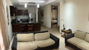 A living room with a sofa and a coffee table, a kitchen area with a refrigerator and a dining table, and a bedroom visible through an open door. - Beachfront Jaco Condos - Fully Equipped - 6 Pax (Jacó)