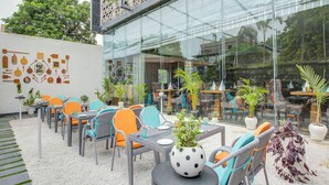 Un espacio al aire libre con sillas y mesas de mimbre, rodeado de plantas en macetas y una pared de vidrio que refleja el interior.