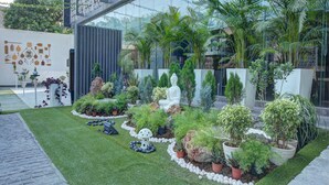 Un jardín bien cuidado con diversas plantas y una estatua de Buda, rodeado por un edificio moderno con amplias ventanas.