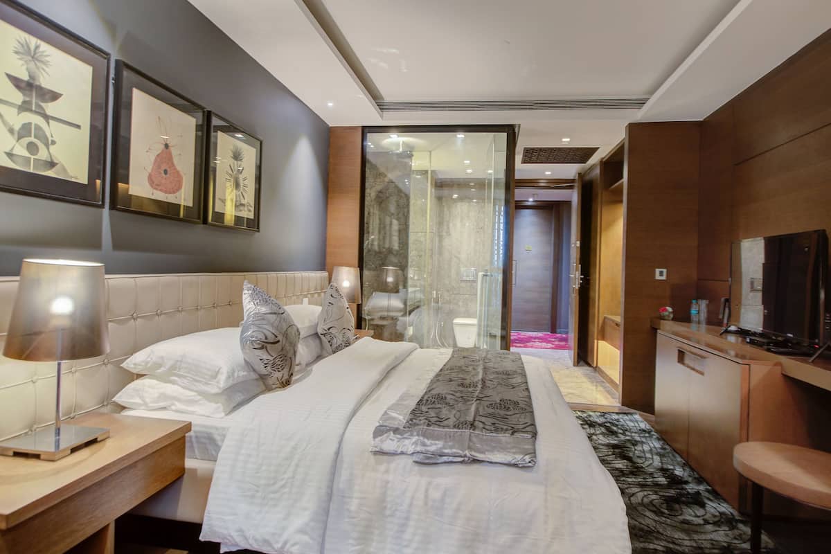 A modern hotel room with a neatly made bed, a bedside table with a lamp, a desk with a chair, and a TV.