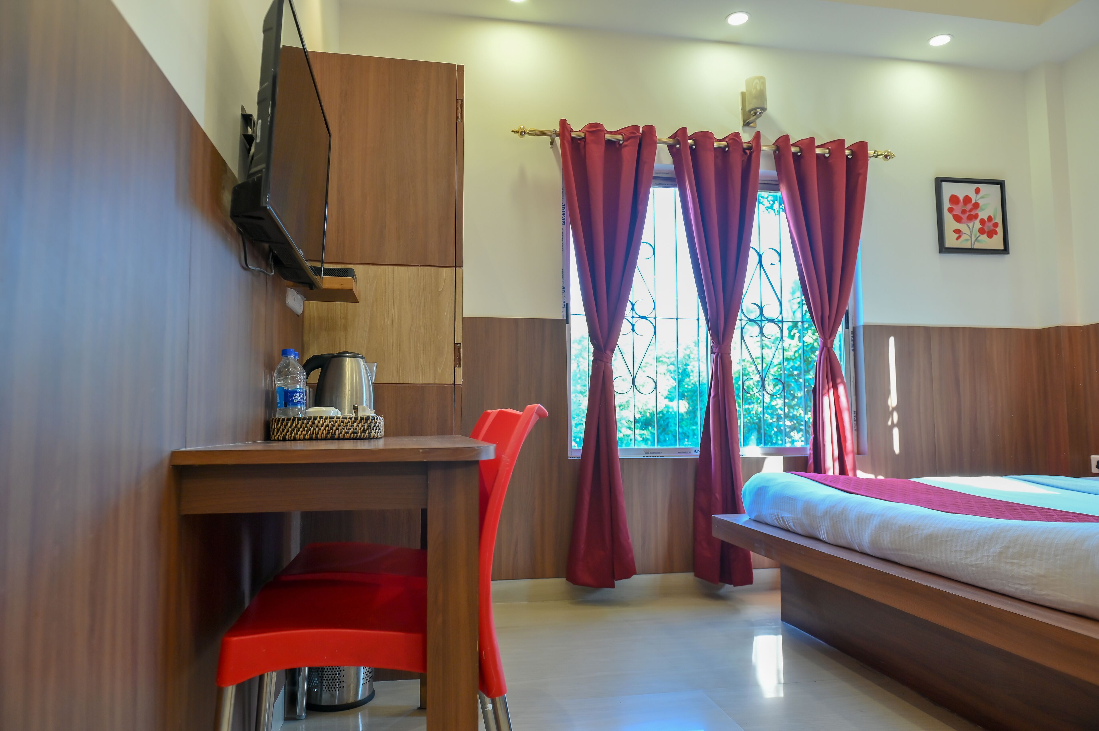A bedroom with a bed, a desk with a chair, a TV mounted on the wall, and a window with curtains.