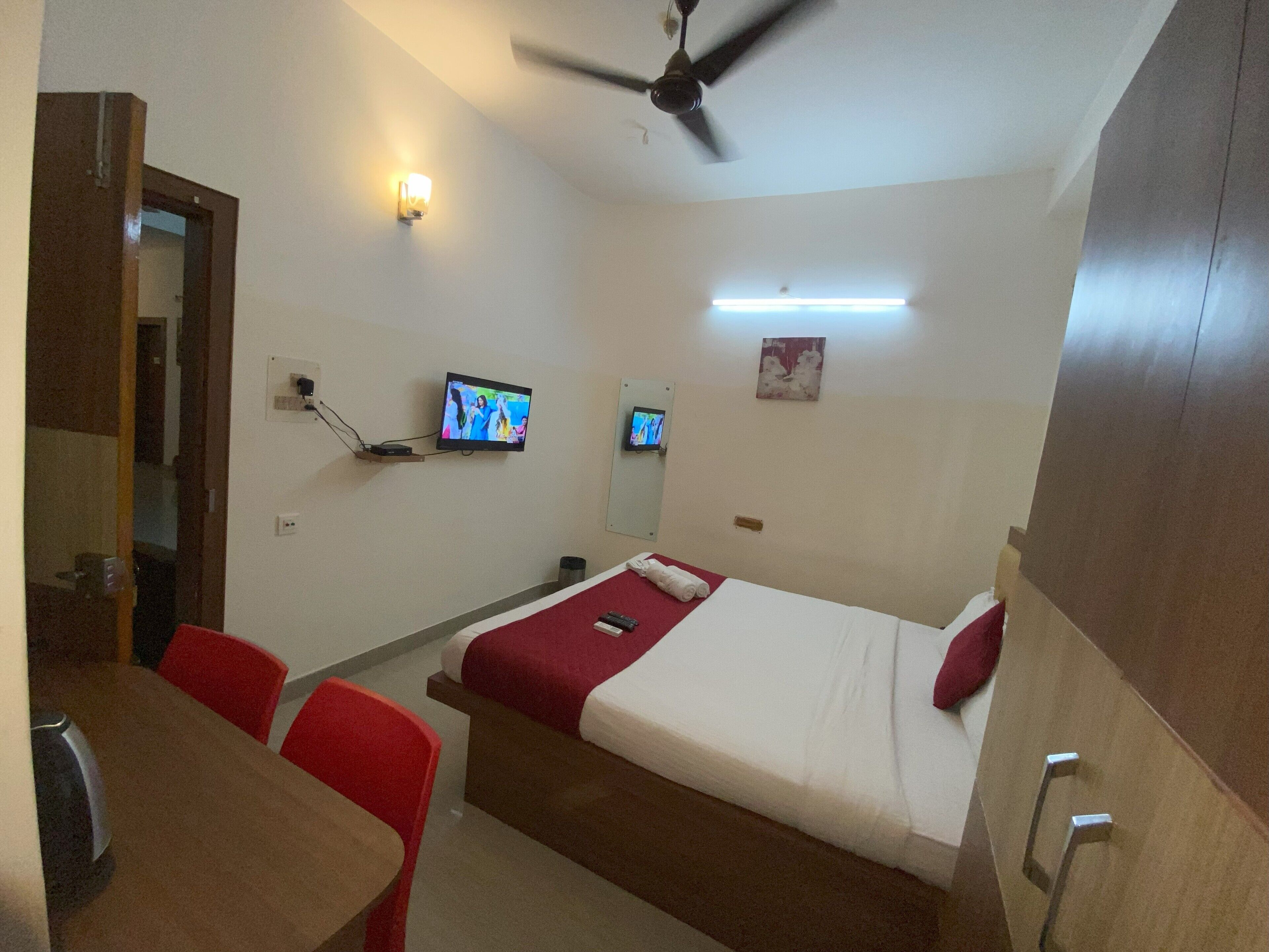 A hotel room with a bed, a desk, two chairs, a ceiling fan, and two wall-mounted TVs.