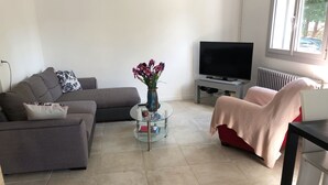Una sala de estar moderna con un sofá gris, una mesa de centro redonda de vidrio, una silla roja con una manta, un televisor de pantalla plana y un radiador.