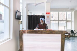 A reception desk with a person seated behind it, a television mounted on the wall, and a window with a view of a building outside. - OYO 43300 Spring Woods Suites (Mohali)