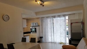 A living room with a beige sofa, a dining table with chairs, a kitchen with a built-in oven, and a clock on the wall. - T3 in Saint Cyprien, 400m from the beach (Saint-Cyprien Plage)