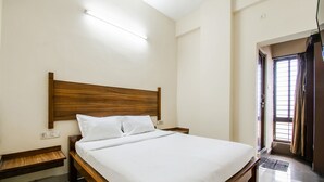 Ein ordentlich bezogenes Bett mit hölzernem Kopfteil in einem Hotelzimmer mit Fliesenboden und einem fensterbelaubten Fenster.