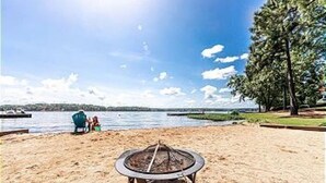 A beach with a fire pit in the foreground, a lake in the background, and a couple sitting on the sand.