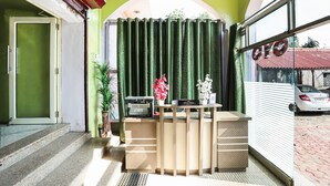 Un mostrador de recepción con una impresora, un letrero que dice "WIZARD" y una planta en maceta. Se ve una cortina verde detrás del mostrador, y un coche es visible a través de la ventana.
