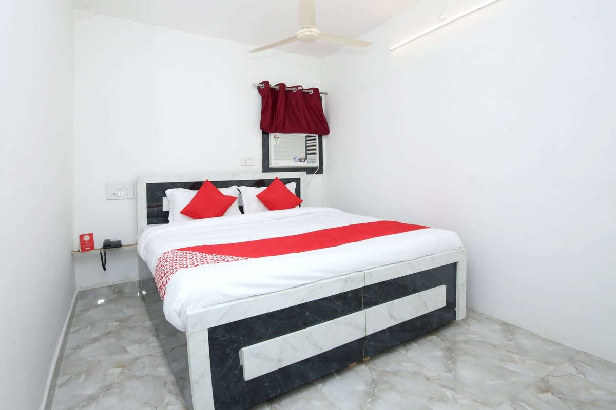 A bedroom with a bed, white walls, a ceiling fan, a window with curtains, and a nightstand with a phone.