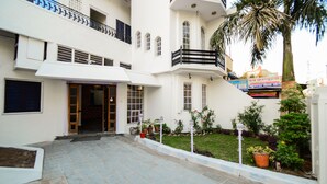 A two-story hotel with a white facade, a balcony on the upper floor, and a well-maintained garden with potted plants and a palm tree.