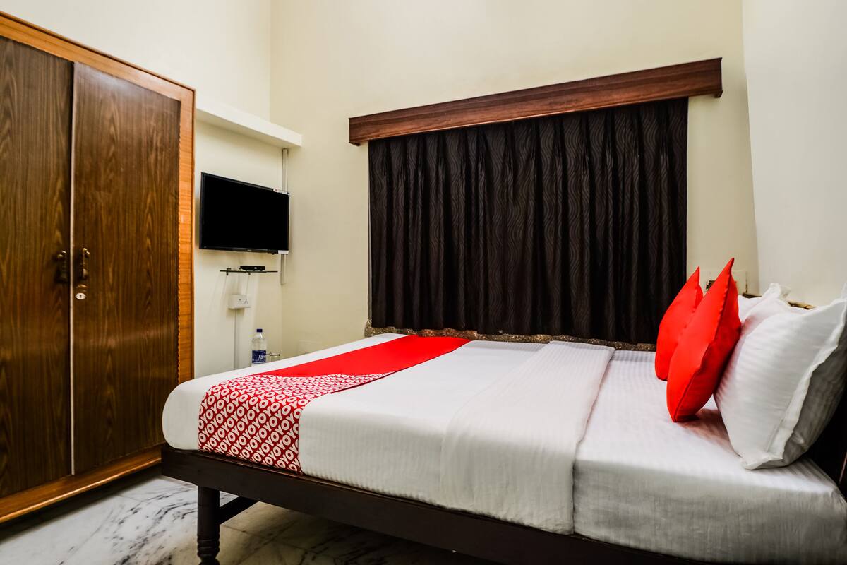 Cama bem arrumada com colcha vermelha e branca, guarda-roupa de madeira e televisão de tela plana na parede.
