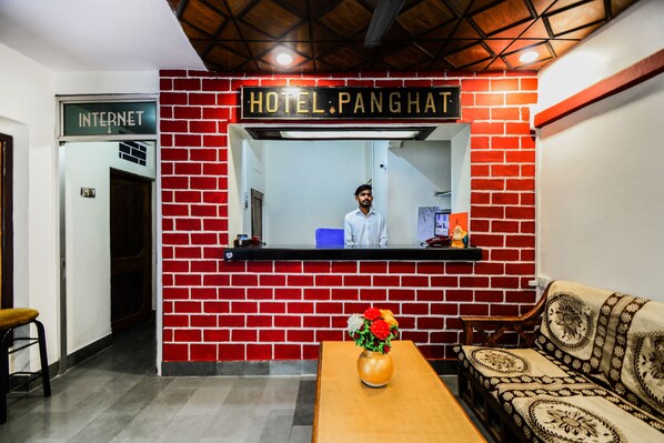 Un área de recepción con una pared de ladrillo rojo, un cartel que dice 'HOTEL PANGHAT', un mostrador de recepción, un sofá y un jarrón con flores.