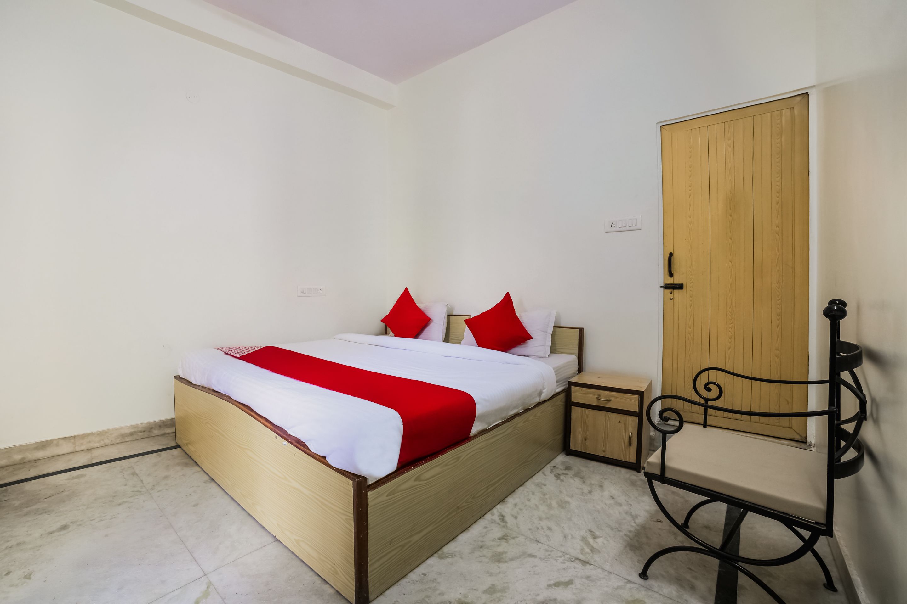 A single bed with a wooden frame, a red and white bedspread, and two red pillows. There is a wooden wardrobe with a black metal chair beside it.