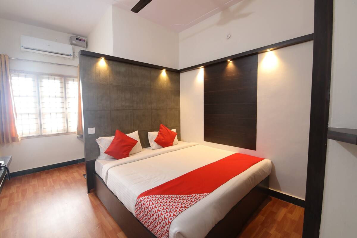 A modern hotel room with a large bed, two red pillows, a red and white patterned throw, and a wooden floor.