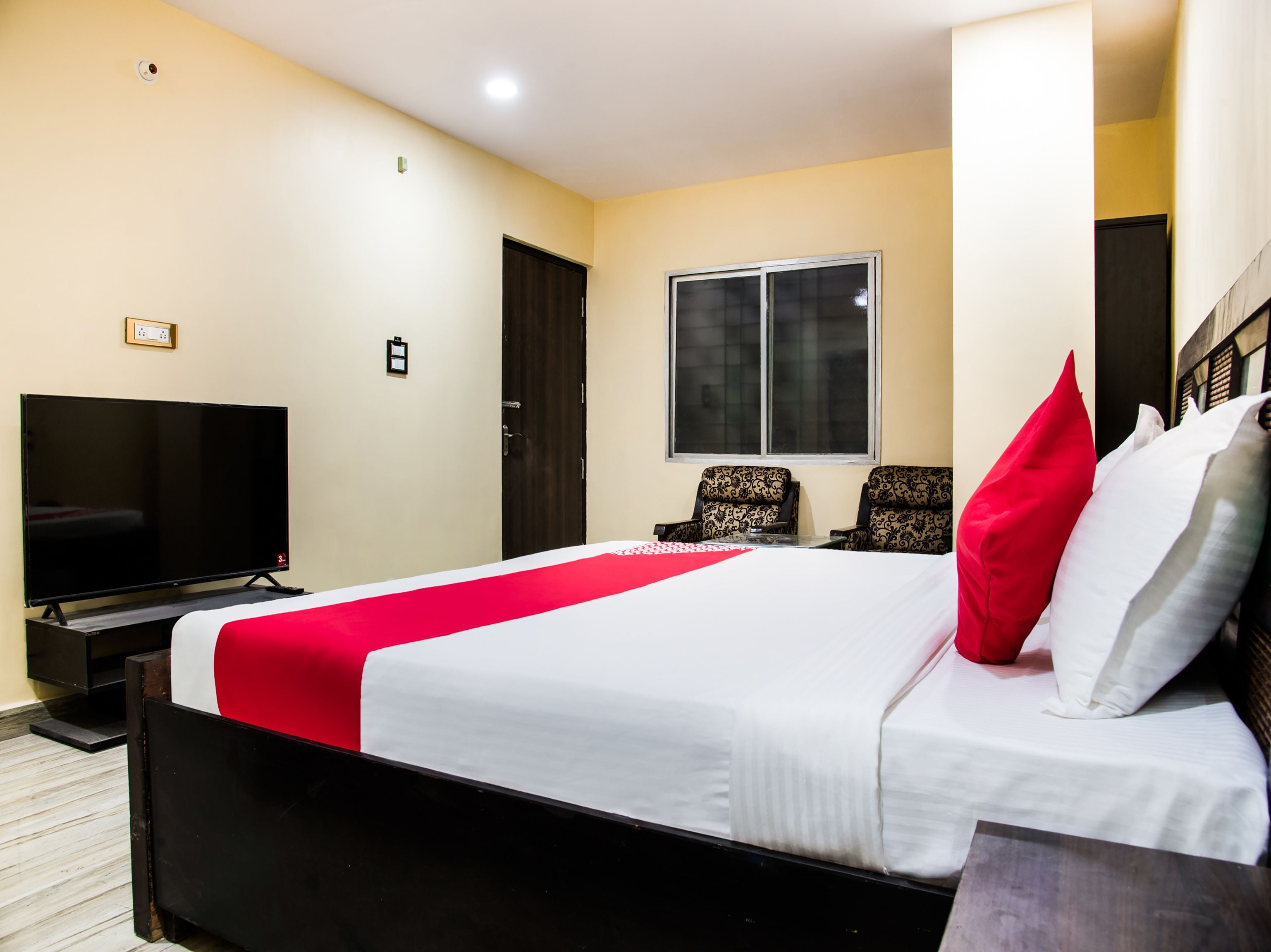A hotel room with a large bed, a television, a window, and two patterned chairs.