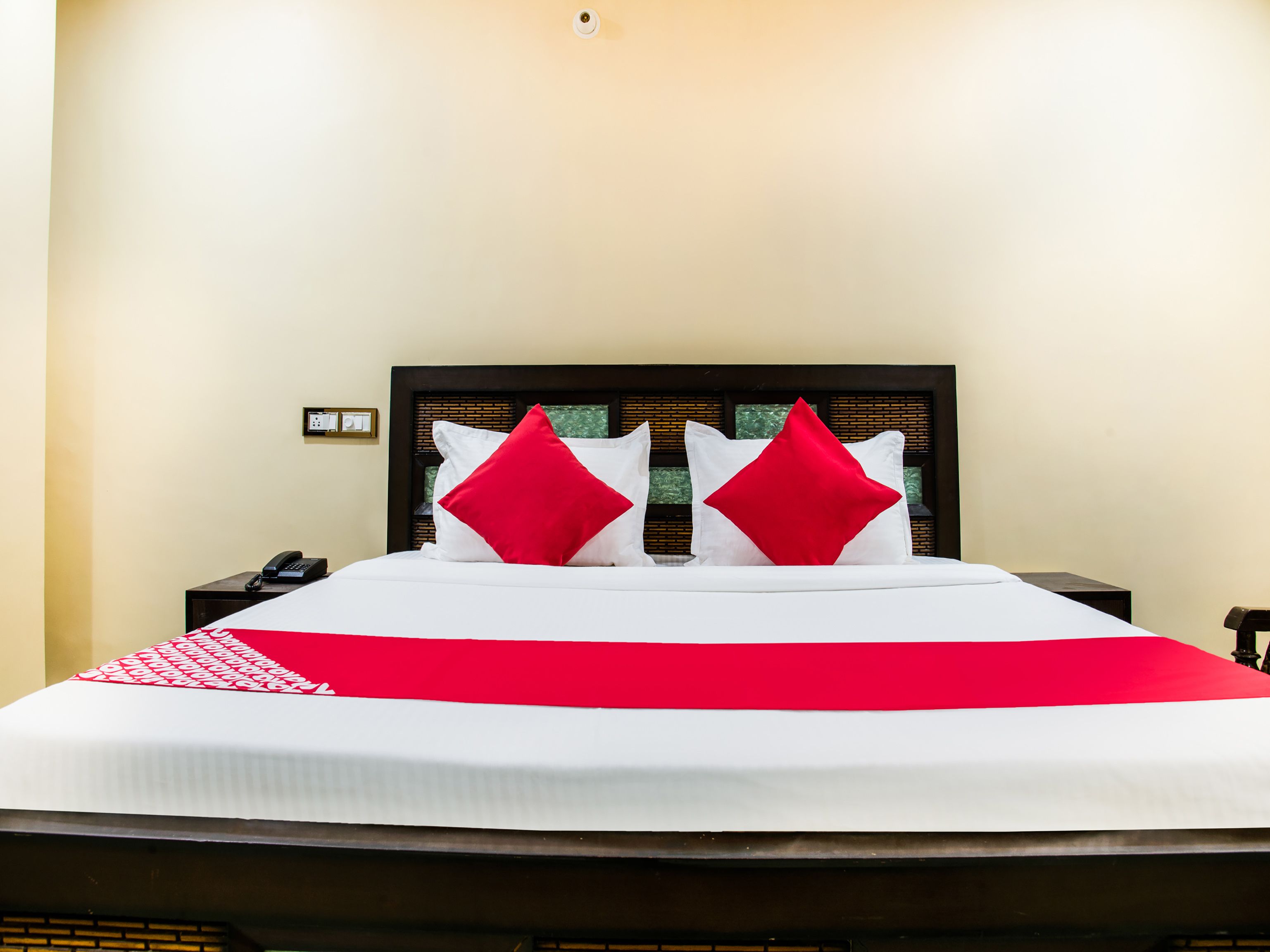 A neatly made bed with white and red bedding, a wooden headboard, and a bedside table with a telephone.