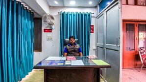 A man sitting at a desk in an office with blue curtains, a fan, and various papers. - OYO Flagship 23420 Patliputra Heritage Guest House (Patna)