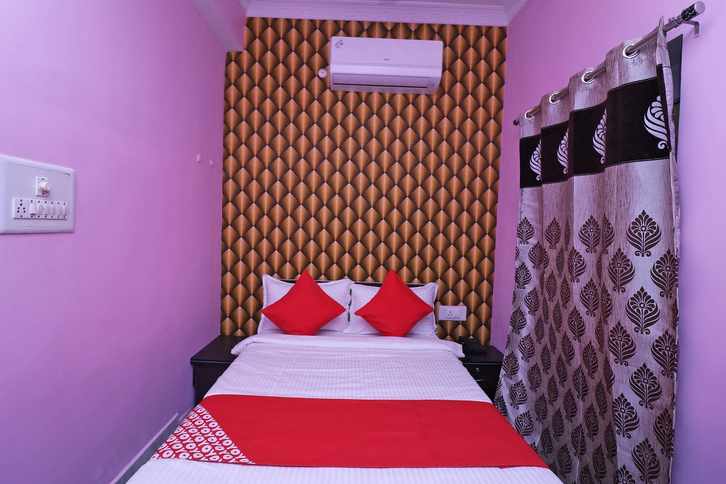Un dormitorio con cabecera estampada, cama con ropa de cama roja y blanca, mesita de noche y cortina con diseño de hojas.