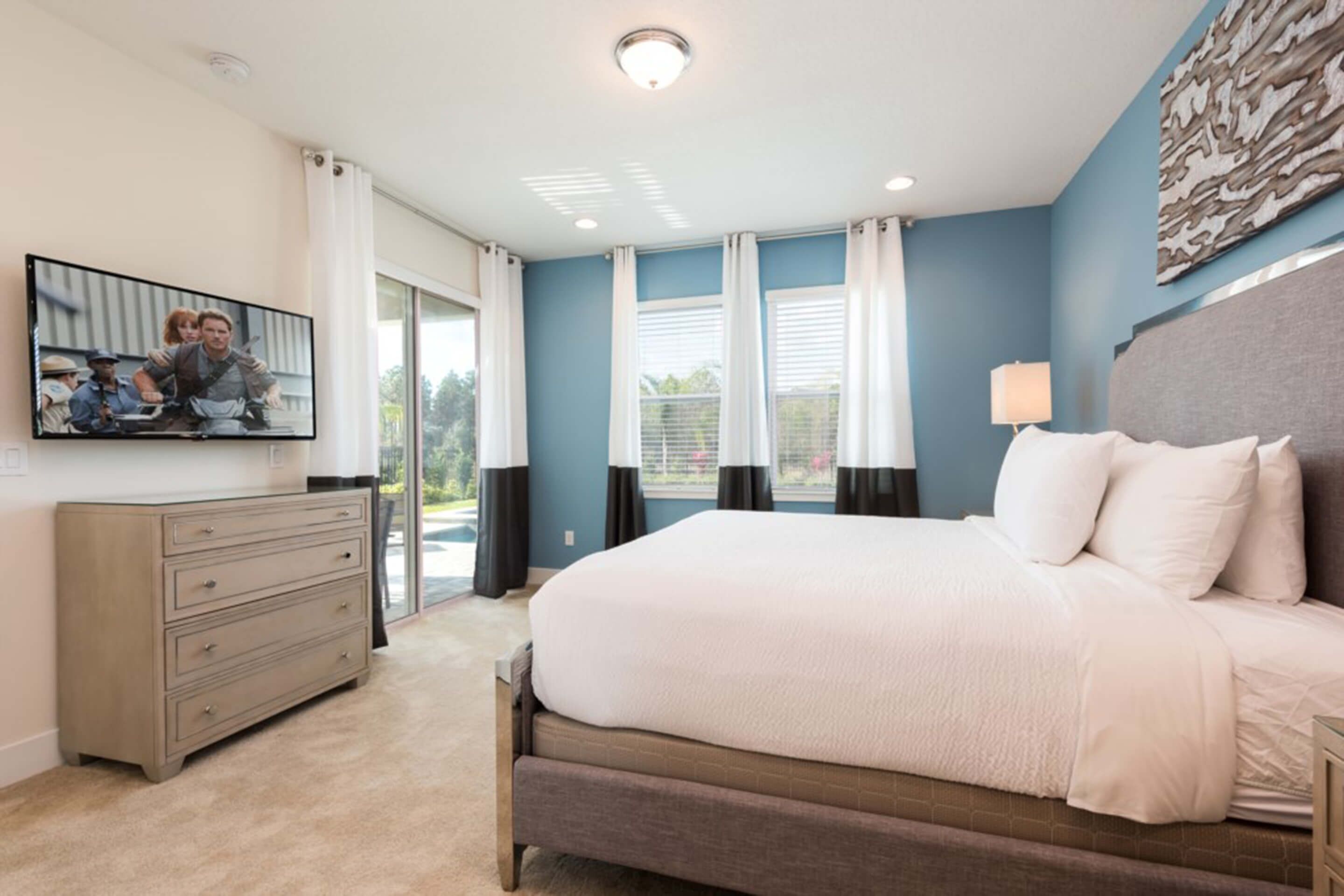 A bedroom with a large bed, a dresser, a TV mounted on the wall, and a window with curtains.