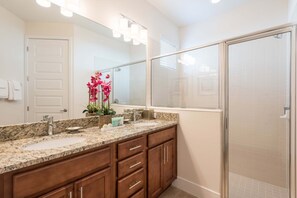 A bathroom with a granite countertop sink, wooden cabinets, a large mirror with lights, and a glass shower enclosure. - Diamond Villa (Kissimmee)