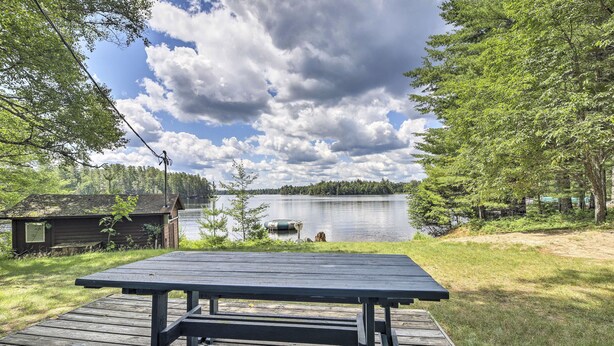 Una terraza de madera con una mesa de picnic que da a un lago, rodeada de árboles y una pequeña cabaña.
