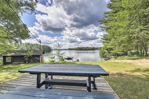 Una terraza de madera con una mesa de picnic dando a un lago, rodeada de árboles y una cabaña pequeña.