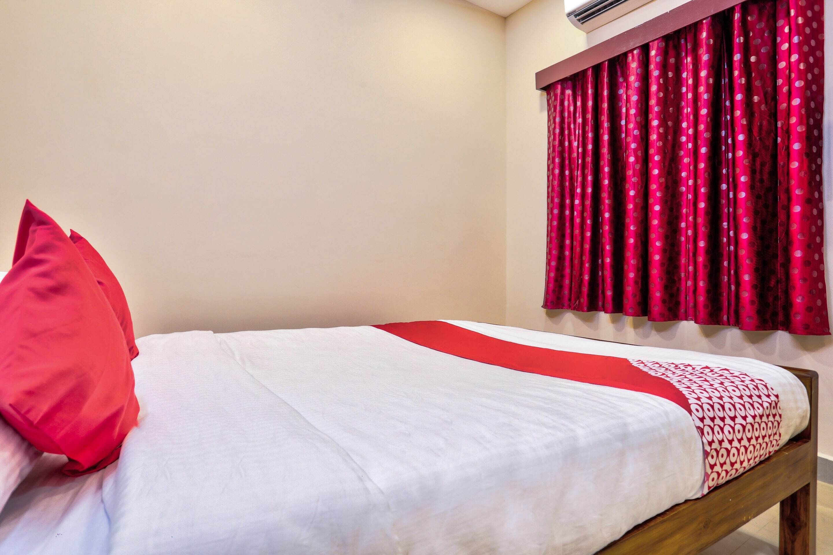A single bed with a white and red bedspread, a red pillow, and a window with red curtains.