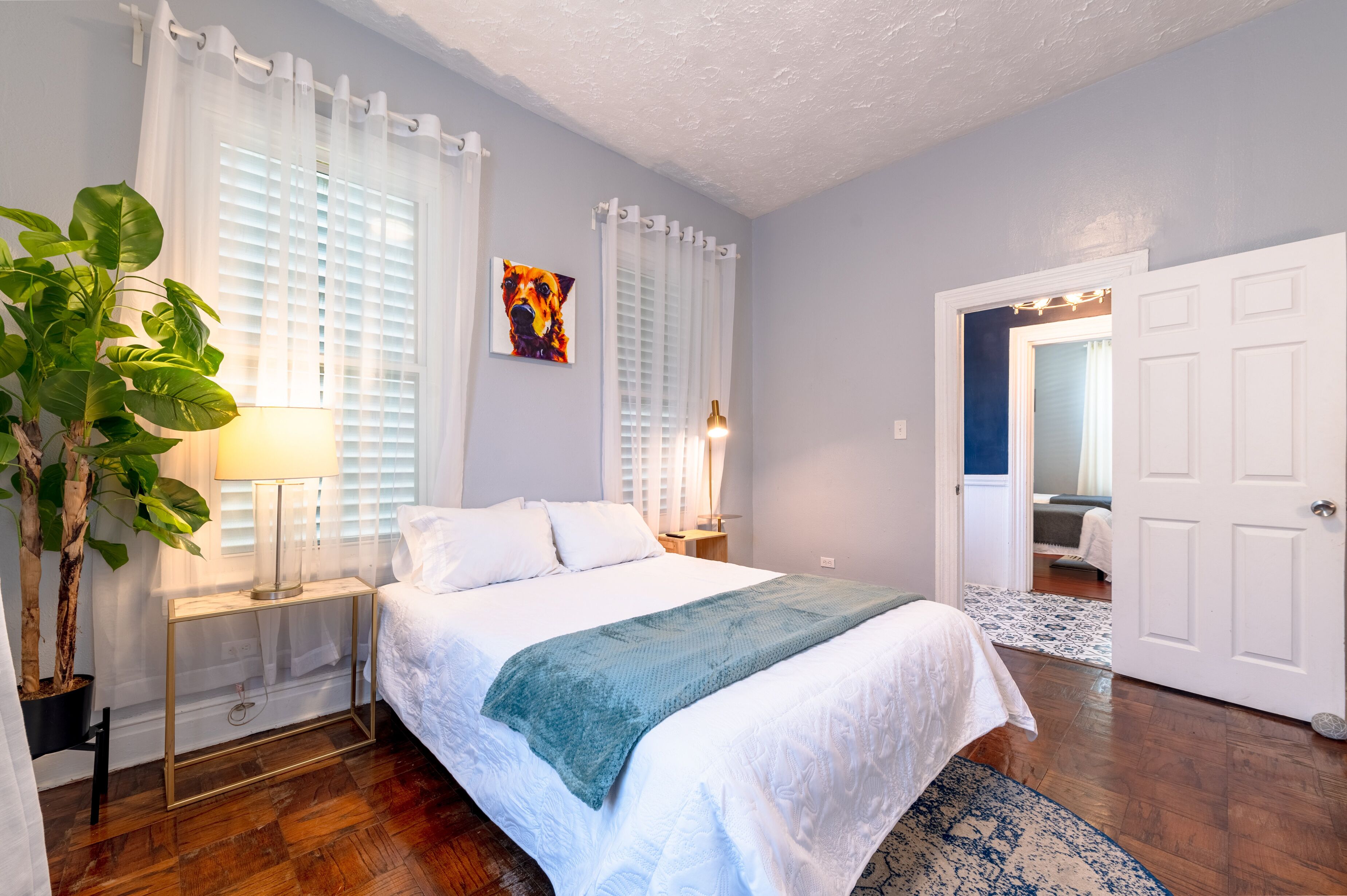 A bedroom with a bed, bedside lamps, a plant, and a door leading to another room.