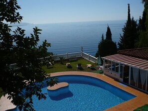 Ein Pool mit Blick auf das Meer, umgeben von einer Sonnenterrasse und einem überdachten Bereich.