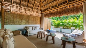 A room with a thatched roof, a sofa, a blue ottoman, and a view of lush greenery outside. - Villa Amalfi (Tulum)