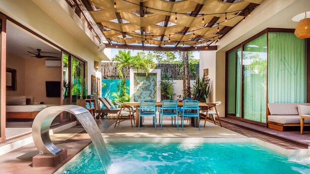 A pool area with a waterfall feature, a dining table with chairs, and a sofa. - Villa Amalfi (Tulum)