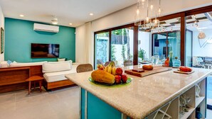 A modern kitchen with a central island, a dining area with a sofa, and a large window providing a view of the outdoors. - Villa Amalfi (Tulum)