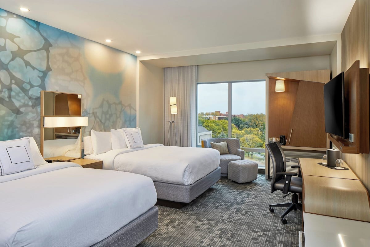 Habitación de hotel con dos camas, un escritorio, una silla, un televisor y un ventanal con vistas a árboles.