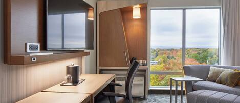 Una habitación de hotel moderna con escritorio, silla y sofá. Tiene un ventanal con vistas al follaje otoñal.