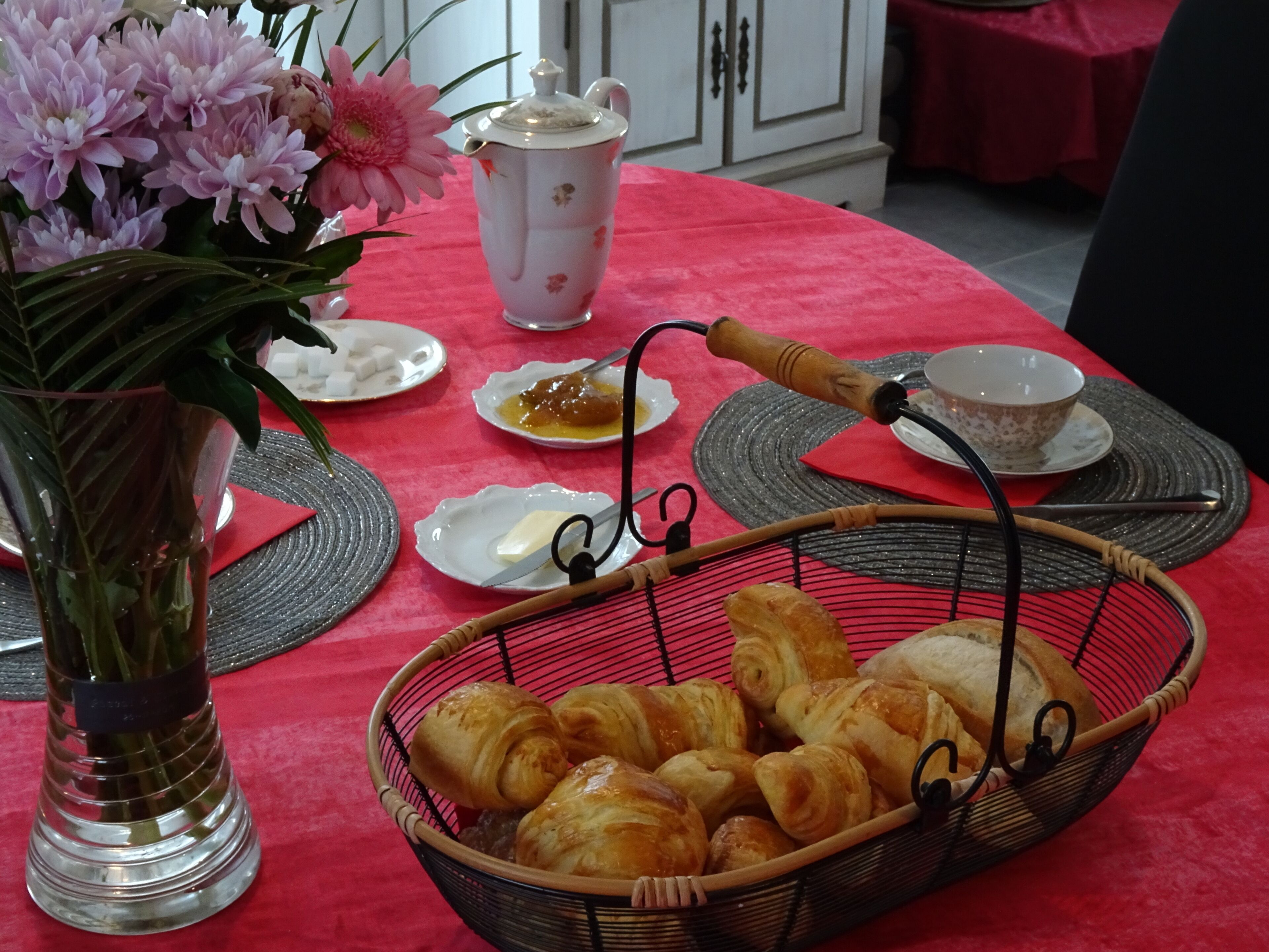 Een eettafel met een rode tafelkleed, een mand met croissants, een theepot en een vaas met bloemen.