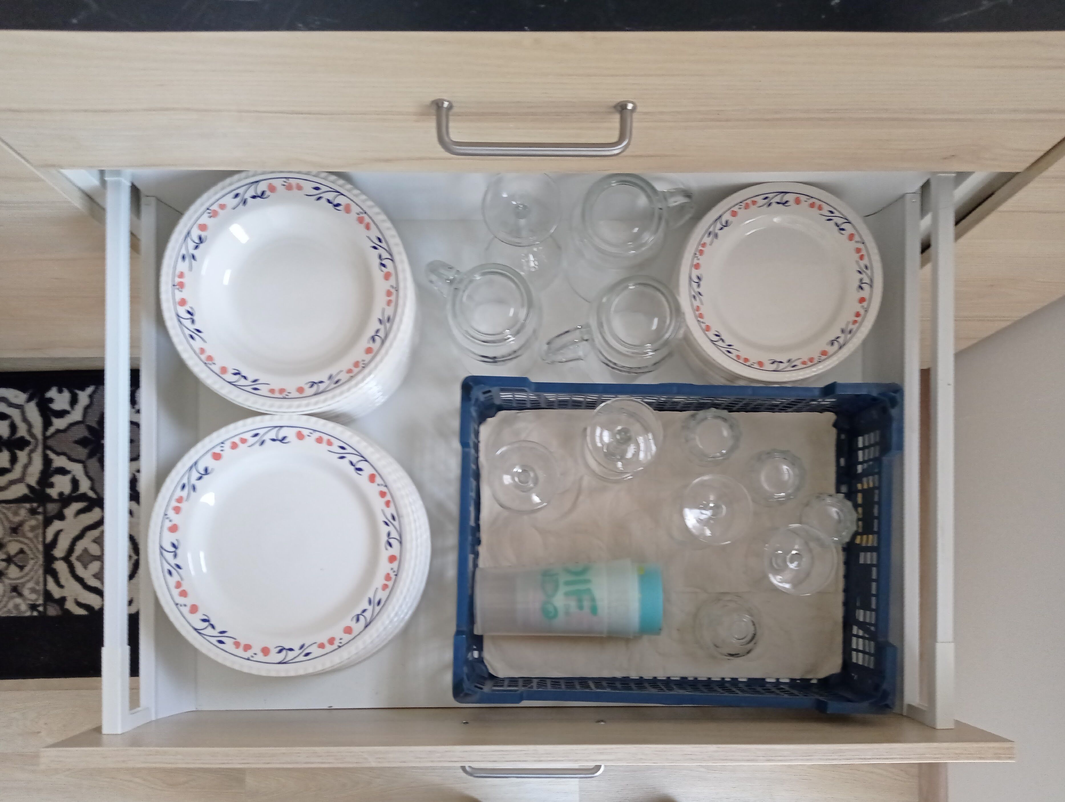 Een lade met witte borden met bloemmotieven, heldere glazen bekers en een blauwe kist met extra drinkglazen.