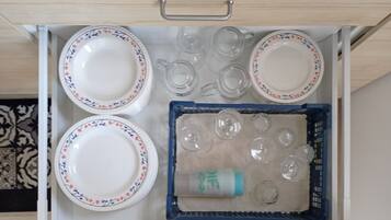 Een lade met witte borden met bloemmotieven, heldere glazen bekers en een blauwe kist met extra drinkglazen.