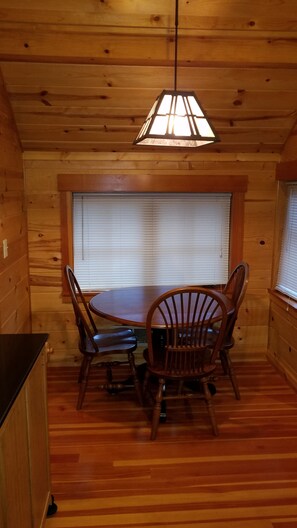Interior de una cabaña de madera con una mesa de comedor y sillas, una lámpara colgante y una ventana con persianas.