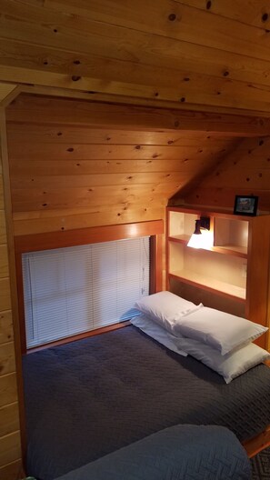 Una habitación pequeña y acogedora con una cama individual, cabecera de madera, ventana con persianas y una estantería con lámpara.