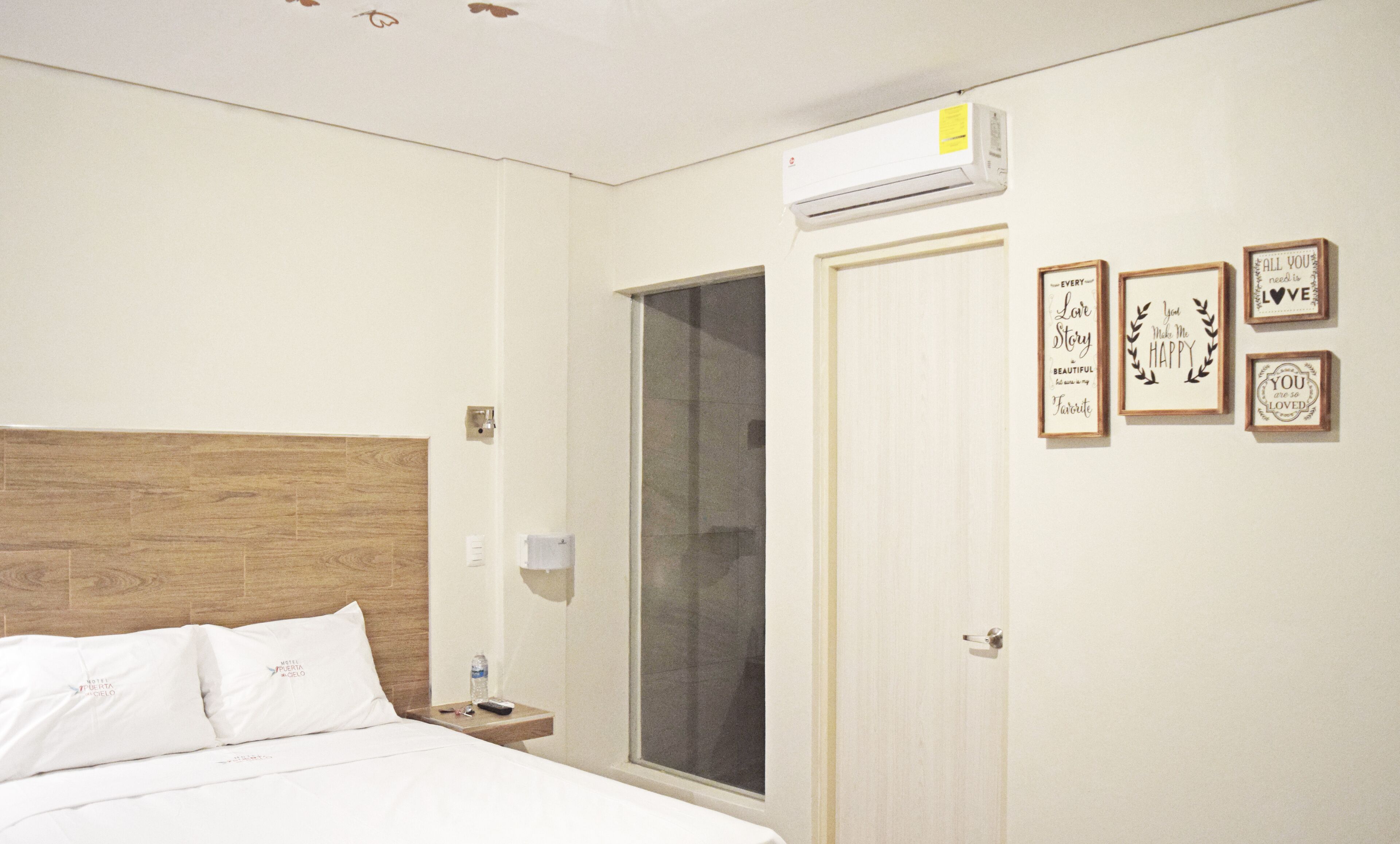 A hotel room with a wooden headboard, a bed with white linens, a nightstand with a remote control, a water bottle, and a door with a window.
