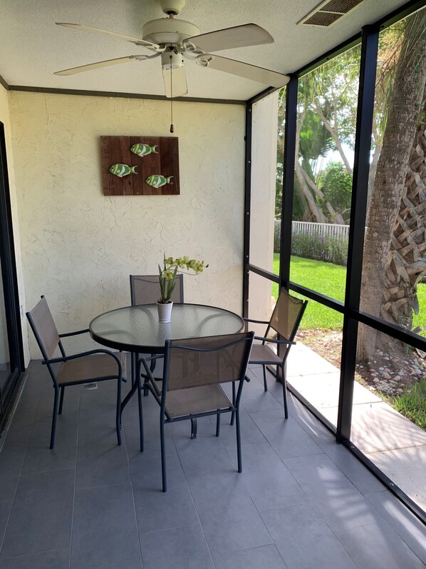 A dining area with a round glass table, four chairs, a ceiling fan, and a view of a garden through large windows. - Contemporary Garden View, Ground Floor Unit, Short Walk to Pool/Beach (Siesta Key)