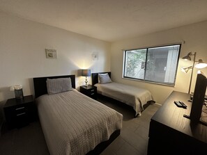 Habitación de hotel con dos camas individuales, un escritorio con lámpara, una mesita de noche y una ventana con persianas.