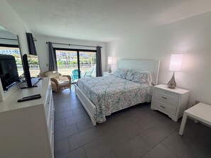 Une chambre d’hôtel équipée d’un lit, d’une télévision, d’un bureau, d’une chaise et d’un balcon offrant une vue sur les bâtiments et la verdure.