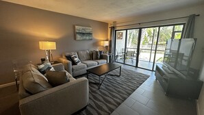 A modern living room with a sofa, armchairs, a coffee table, and a TV. There is a sliding glass door leading to an outdoor area with a table and chairs. - Contemporary Garden View, Ground Floor Unit, Short Walk to Pool/Beach (Siesta Key)