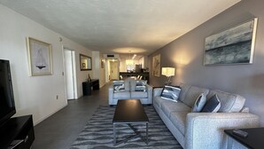 A modern living room with a grey sofa, a coffee table, and a TV. There is a kitchen in the background. - Contemporary Garden View, Ground Floor Unit, Short Walk to Pool/Beach (Siesta Key)