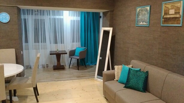 Un salón con sofá, una mesa y una silla. Hay una puerta y una ventana con cortinas.